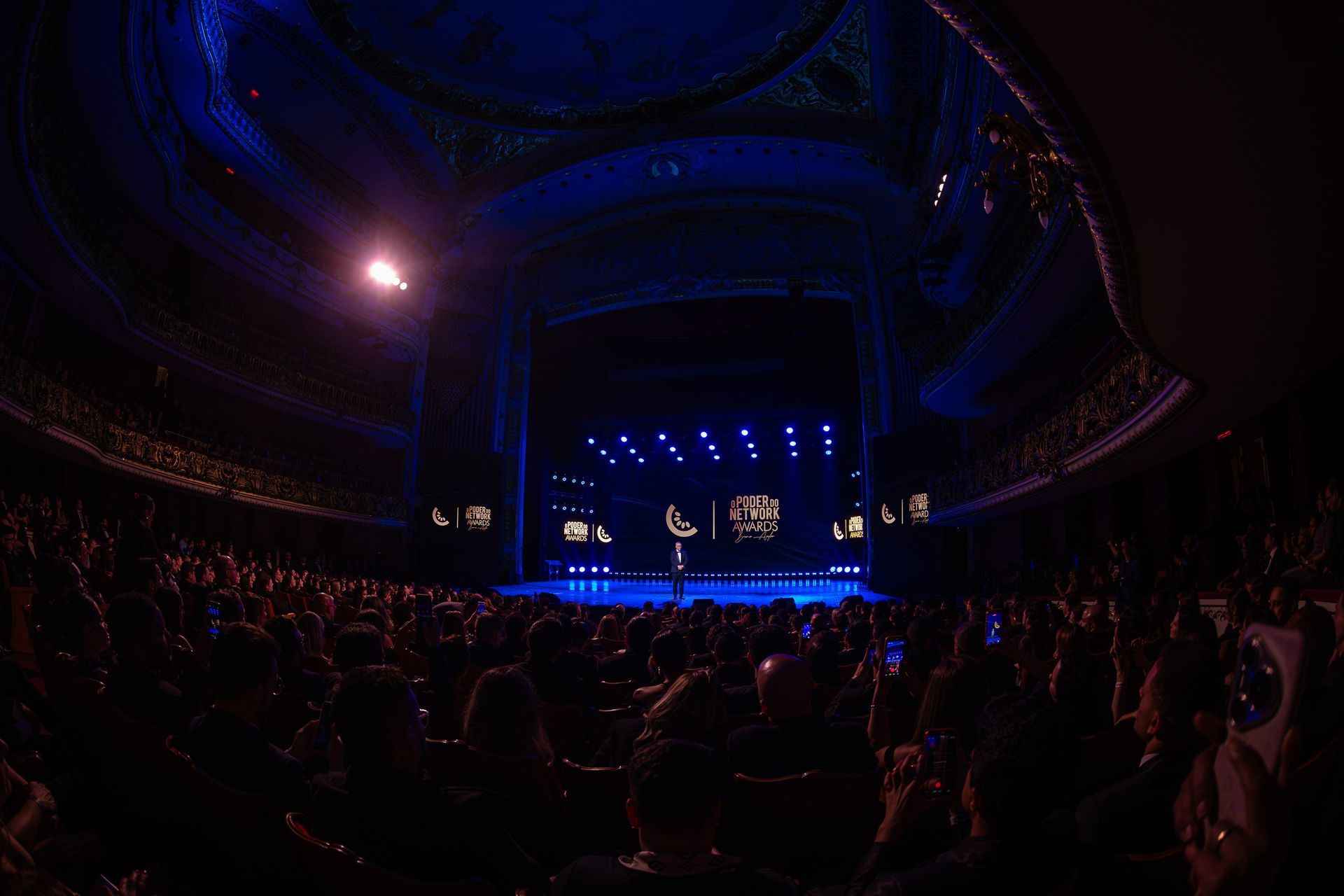 Theatro Municipal lotado durante o Poder do Network Awards, com o palco iluminado em azul e uma figura solitária no centro